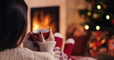Over the shoulder young woman relaxing near fireplace with cup of tee in cozy christmas woolen socks with decorated xmas tree in background