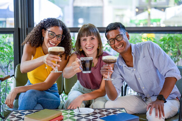 three young millennials toasting with shaken coffees, looking at camera and smiling, people having...