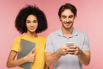 cheerful man chatting on smartphone near latin woman holding laptop isolated on pink.