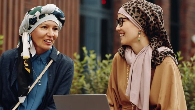Medium close-up of Biracial mature woman wearing turban talking to Muslim Middle Eastern female colleague, who holding portable computer, sitting outdoors