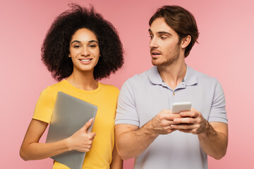 happy hispanic woman with laptop looking at camera near man using cellphone isolated on pink.