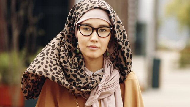 Chest-up of young beautiful Middle Eastern woman wearing hijab and eyeglasses, posing and looking on camera outdoors