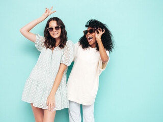 Two young beautiful smiling international hipster female in trendy summer clothes. Sexy carefree women posing near blue wall in studio. Positive models having fun. Concept of friendship. In sunglasses