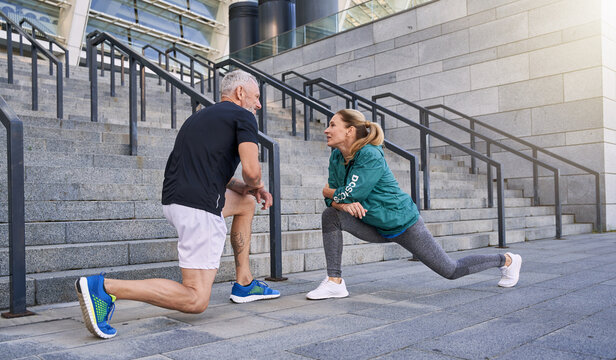 Full length shot of sportive middle aged couple, man and woman stretching legs while exercising together outdoors on a sunny day