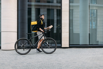 Side view of a male courier walking with a bicycle holding a package. Delivery man with thermal backpack at an office building. - obrazy, fototapety, plakaty