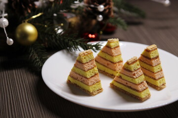 Christmas tree cookies on a white plate, a Christmas tree branch decorated with red and yellow balls on a dark wooden background. New Year's still life with cookies.