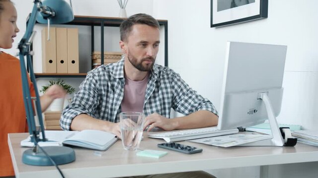 Hyperactive child is bothering annoyed father who is working from home using computer and talking on mobile phone sitting at desk in light room