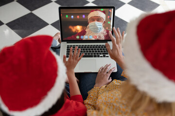Caucasian mother and child in santa hat on christmas laptop video call with santa claus in face mask