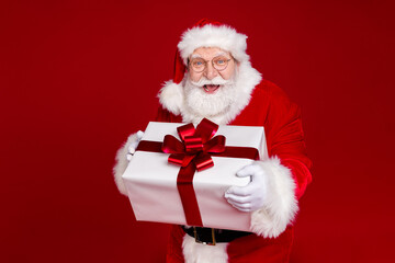 Photo of positive excited retired man wear santa claus costume spectacles smiling holding present box isolated red color background