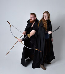 Full length  portrait of red haired  couple, man and woman wearing medieval viking inspired fantasy costumes, standing fighting pose holding  archery bow and arrow and long sword weapons, isolated on 