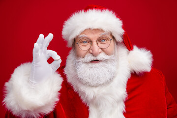 Photo of funny excited retired man wear santa claus costume spectacles smiling showing okey sign isolated red color background