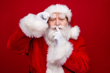 Photo of serious confident retired man wear santa claus costume spectacles asking keep secret finger lips isolated red color background