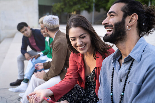 Group of young friends hanging out in the city. Multiracial people sitting by the street and having fun and couple laughing. 