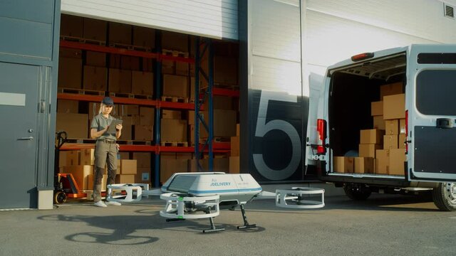 Storage Facility Female Employee Uses Tablet Computer to Send Autonomous Flying Delivery Drone with a Parcel. Futuristic Parcel Drone Takes Off on Flight from Warehouse to Client Waiting for Package