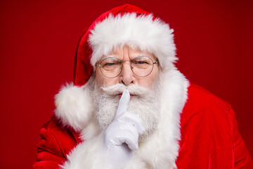 Photo of serious confident retired man wear santa claus costume spectacles fingers lips asking keep silence isolated red color background