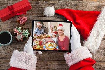 Santa claus holding tablet, making christmas video call with smiling family at dinner table