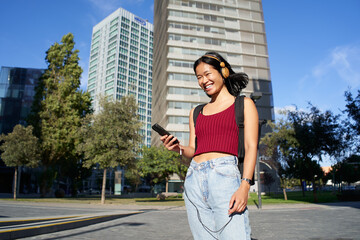 Portrait of cheerful young asian woman out on the city street listening to music or watching video on her mobile phone. Looking at the camera. - obrazy, fototapety, plakaty