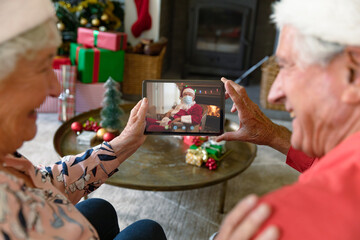 Caucasian senior couple on christmas video call on tablet with santa claus in face mask