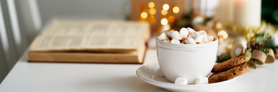 Long banner Christmas light gray background with copy space. Reading book on cozy winter evening with candles, cocoa hot chocolate with marshmallows and cookies. Side view of table, chair in home