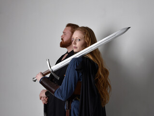 Full length  portrait of red haired  couple, man and woman wearing medieval viking inspired fantasy costumes, standing fighting pose holding  sword weapons, isolated on white  studio background.
