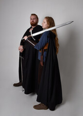 Full length  portrait of red haired  couple, man and woman wearing medieval viking inspired fantasy costumes, standing fighting pose holding  sword weapons, isolated on white  studio background.