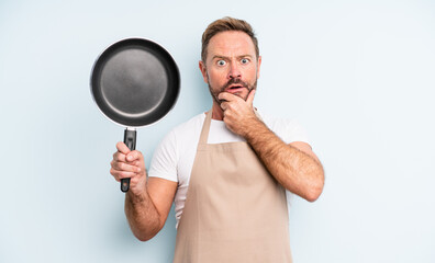 middle age handsome man with mouth and eyes wide open and hand on chin. frying pan concept