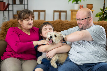 The son is hugging the dog tightly. The parents are sitting next to the son on the sofa in the...