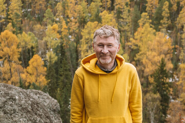 An authentic portrait of a smiling unshaven male traveler in a yellow hoodie on top of a mountain, against the background of an autumn forest. Selective focus. Close-up. Sustainable environment. - Powered by Adobe