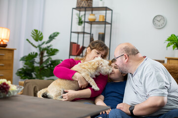 A dog licks the face of a boy who is sitting on the living room sofa with his parents.