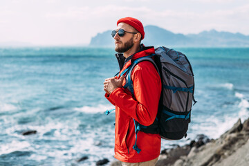 A bearded traveler with a backpack on the background of the sea. Portrait of a traveler in red...