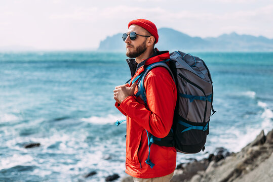 A bearded traveler with a backpack on the background of the sea. Portrait of a traveler in red clothes and sunglasses. A tourist with a backpack stands against the background of the sea. Copy space