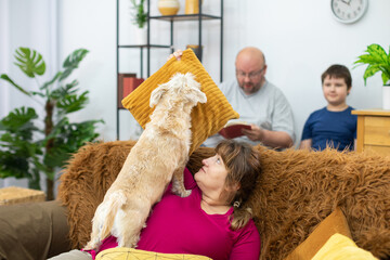 The mother is playing with the dog on the sofa and the father is reading a book to his son.