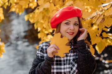 Beautiful caucasian adult young woman in a red beret hat stands near a pond in autumn during leaf fall and smiles emotionally