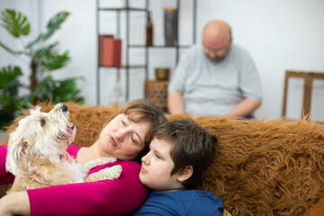 Mother and son are sitting on the sofa and holding a dog in their arms. The father is reading a book.