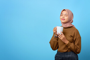 Beautiful young Asian woman holding mug and drinking tea on blue background