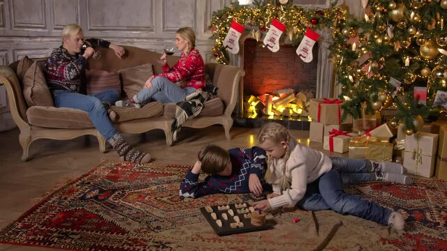 The family (mother, father, son and daughter) spend the winter at home. The apartment is decorated for the New Year or Christmas holidays. Children play a board games, parents drink wine from glasses