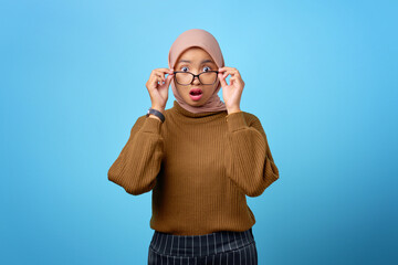 Surprised young Asian woman touching glasses and looking camera on blue background