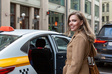 A beautiful young woman gets into a yellow taxi in the city. Smiling blonde in a beige coat. Travel...