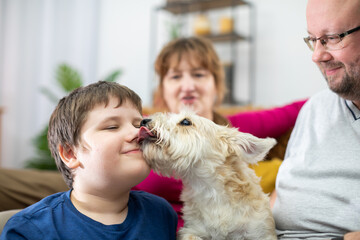 A dog licks the face of a boy who is sitting on the living room sofa with his parents.