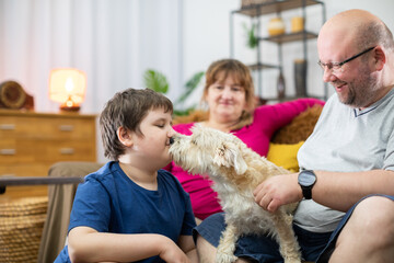 A dog licks the face of a boy who is sitting on the living room sofa with his parents.
