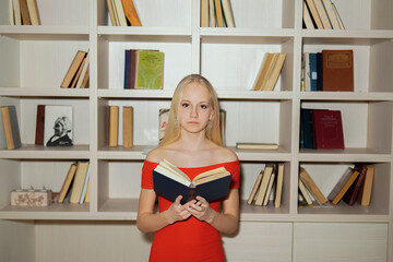 little girl teenager blonde in a red dress reading books on the background of bookshelves.