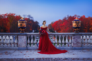 A beautiful girl in a long red ball gown standing on the balcony against the background of an autumn park in the twilight light.