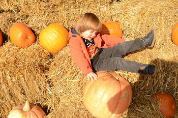 A boy, three years old, at an autumn festival among pumpkins and haystacks. Festive decorations, lots of beautiful pumpkins, haystacks. Halloween. Good mood. The child is wearing an orange jacket