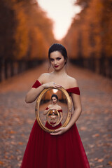 A young girl in a red ball gown holding in her hands a mirror in a vintage frame that reflects...