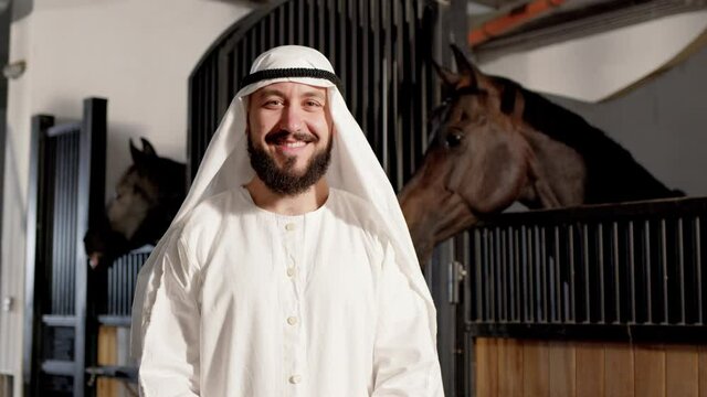 Waist up portrait view of the smiling bearded arabian sheikh smiling to the camera while standing at the stable with his horses at the background. Hobbies for rich people concept