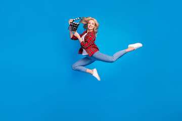 Fototapeta na wymiar Photo of adorable funky young lady wear plaid shirt smiling humping high holding clapperboard isolated blue color background