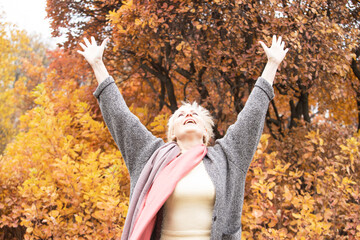 Happy senior woman with hands up standing in autumn park. Adult woman smiling looks up with raised hands. Yellow trees on background. Retirement, elderly health, life insurance, free breathing concept