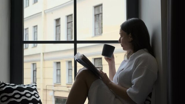 Caucasian Woman in White Bathrobe Drinking Coffee and Reading Book while Sitting on a Windowsill of Hotle Room, Looking Through a Window with a City View