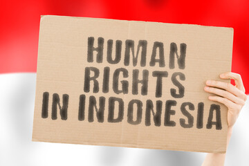 The phrase " Human rights in Indonesia " on a banner in men's hand with blurred Indonesian flag on the background. Dictatorship. Equality. Freedom for people. Restrictions for civil citizens