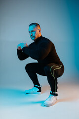 Stylish brutal man doing squat exercises. Athlete workout trending neon blue background. Black...